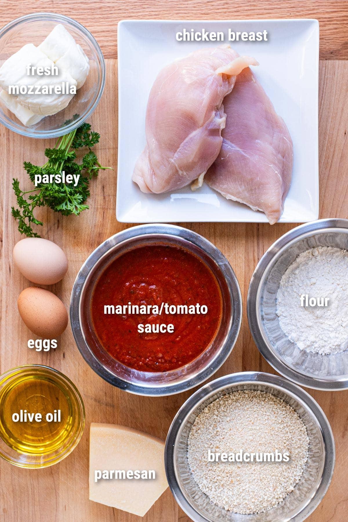 Ingredients for chicken parmesan calzones laid out on a wooden table: chicken breasts, mozzarella cheese, tomato sauce, pizza dough, parmesan cheese, eggs, and breadcrumbs
