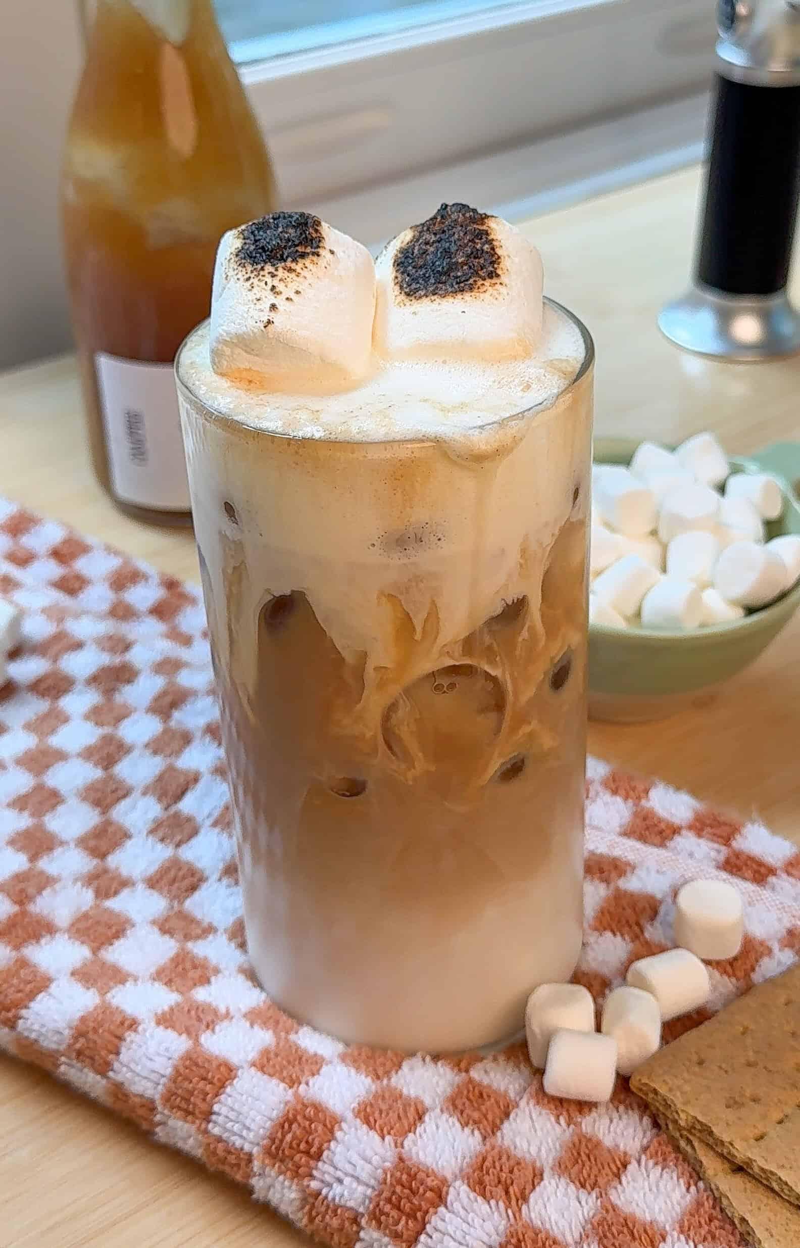 toasted marshmallow chai latte in a clear glass mug, with toasted marshmallows on a wooden skewer resting on the rim