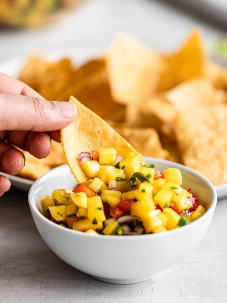 a close-up shot of a person scooping mango salsa onto a tortilla chip, with a blurred beach background