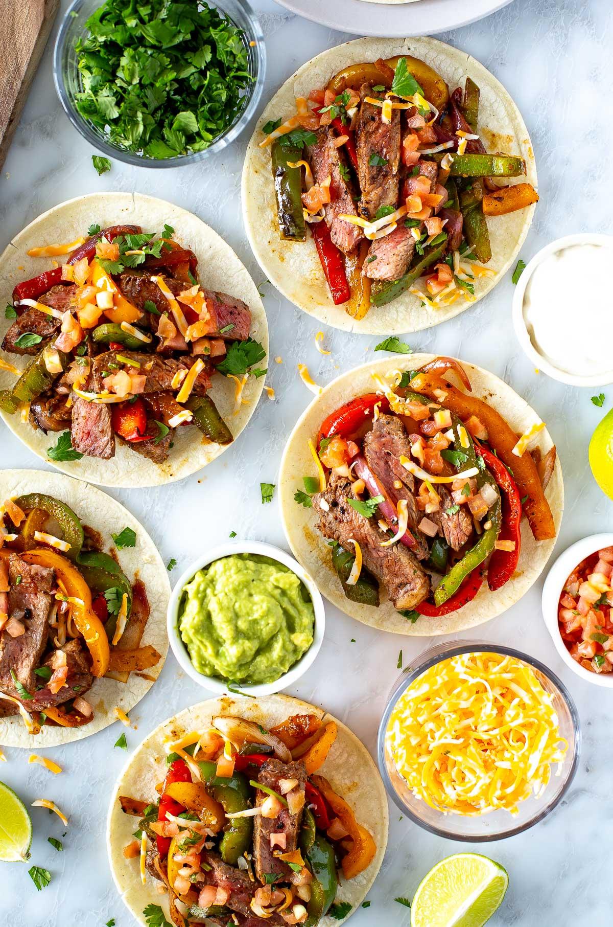 a variety of fajita toppings in small bowls: guacamole, sour cream, salsa, shredded cheese, and cilantro