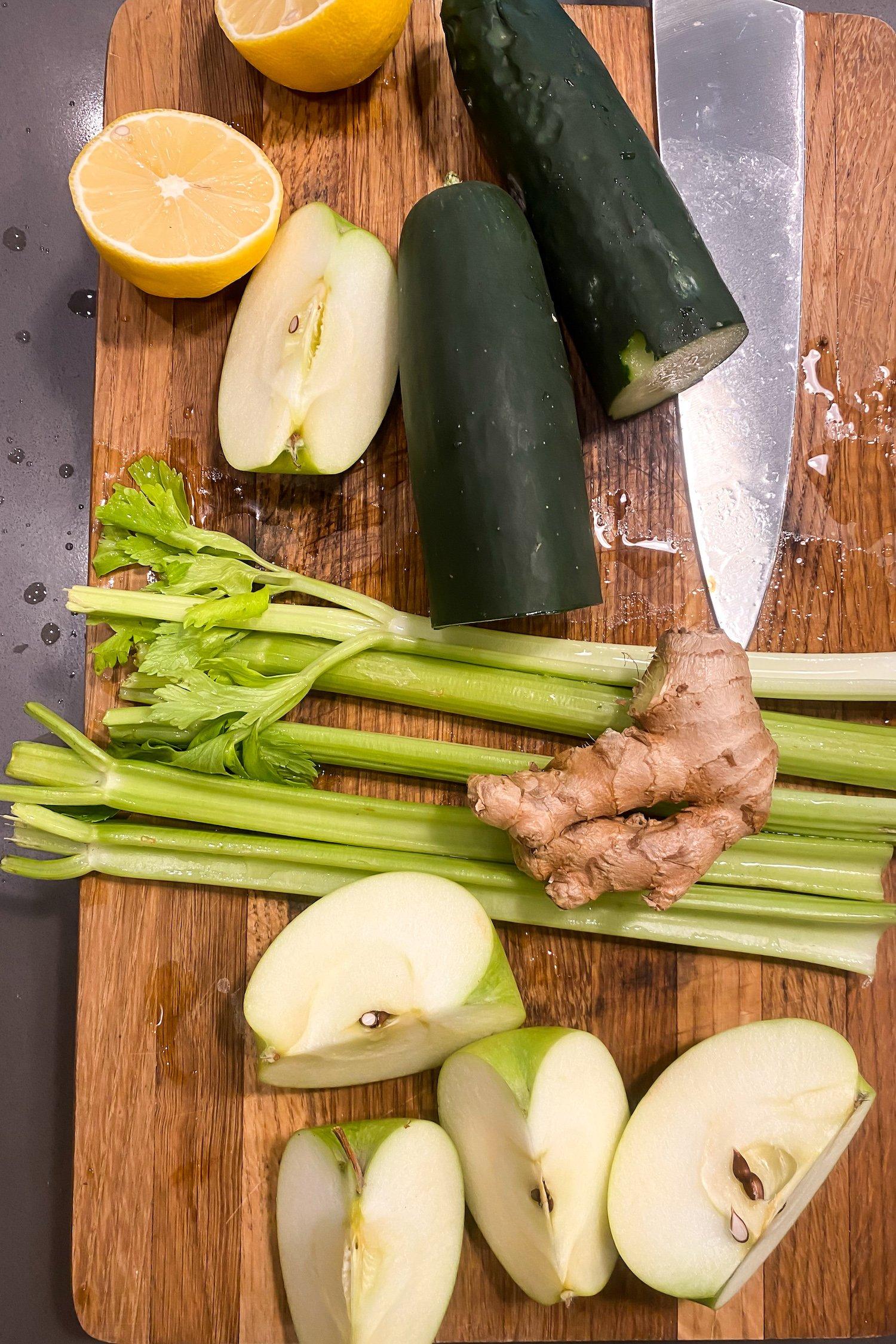 overhead shot of various ingredients for green juice, including apples, ginger, spinach, kale, lemon, and celery, arranged artfully on a wooden cutting board