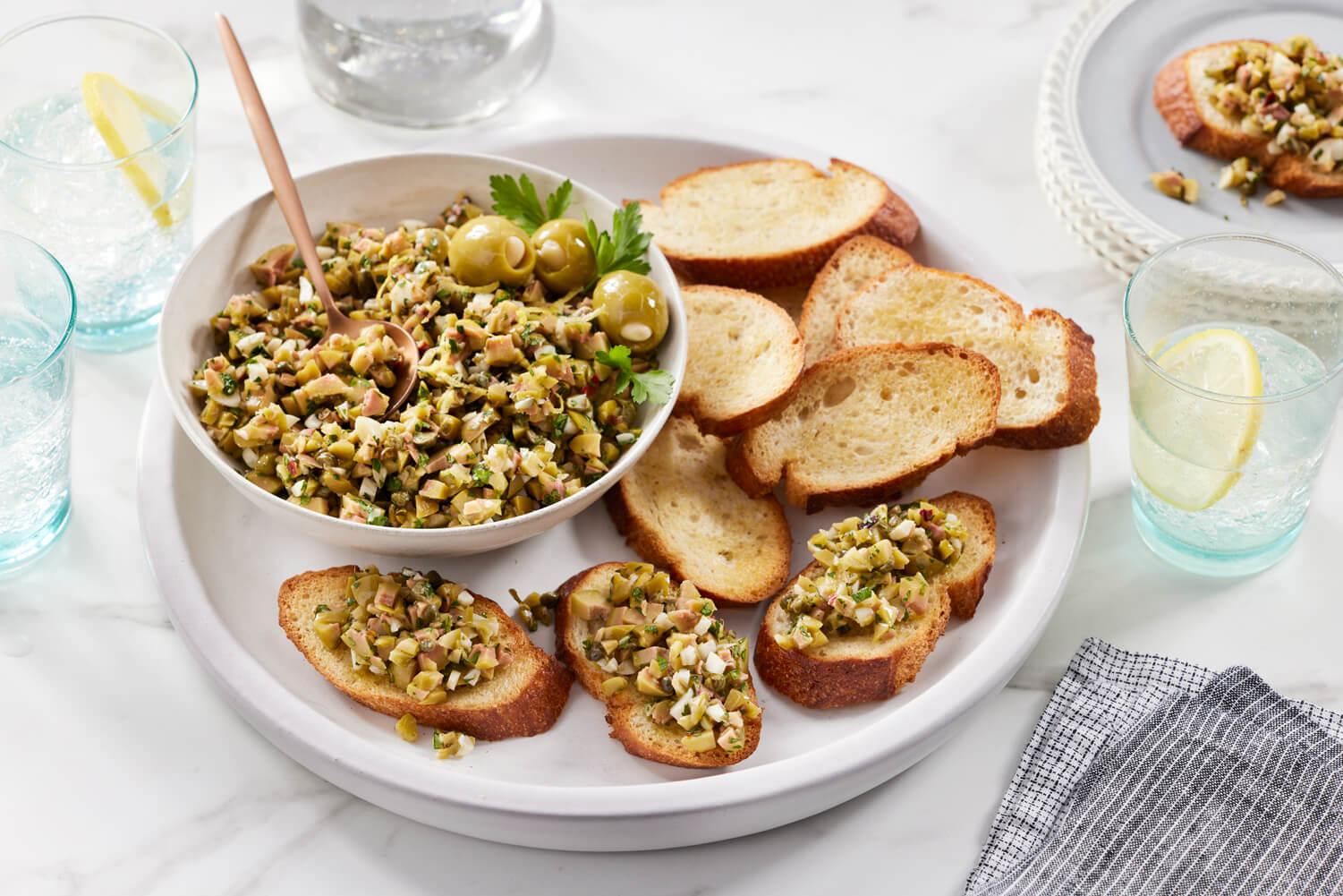 overhead shot of olive tapenade in a rustic bowl surrounded by crostini and fresh vegetables