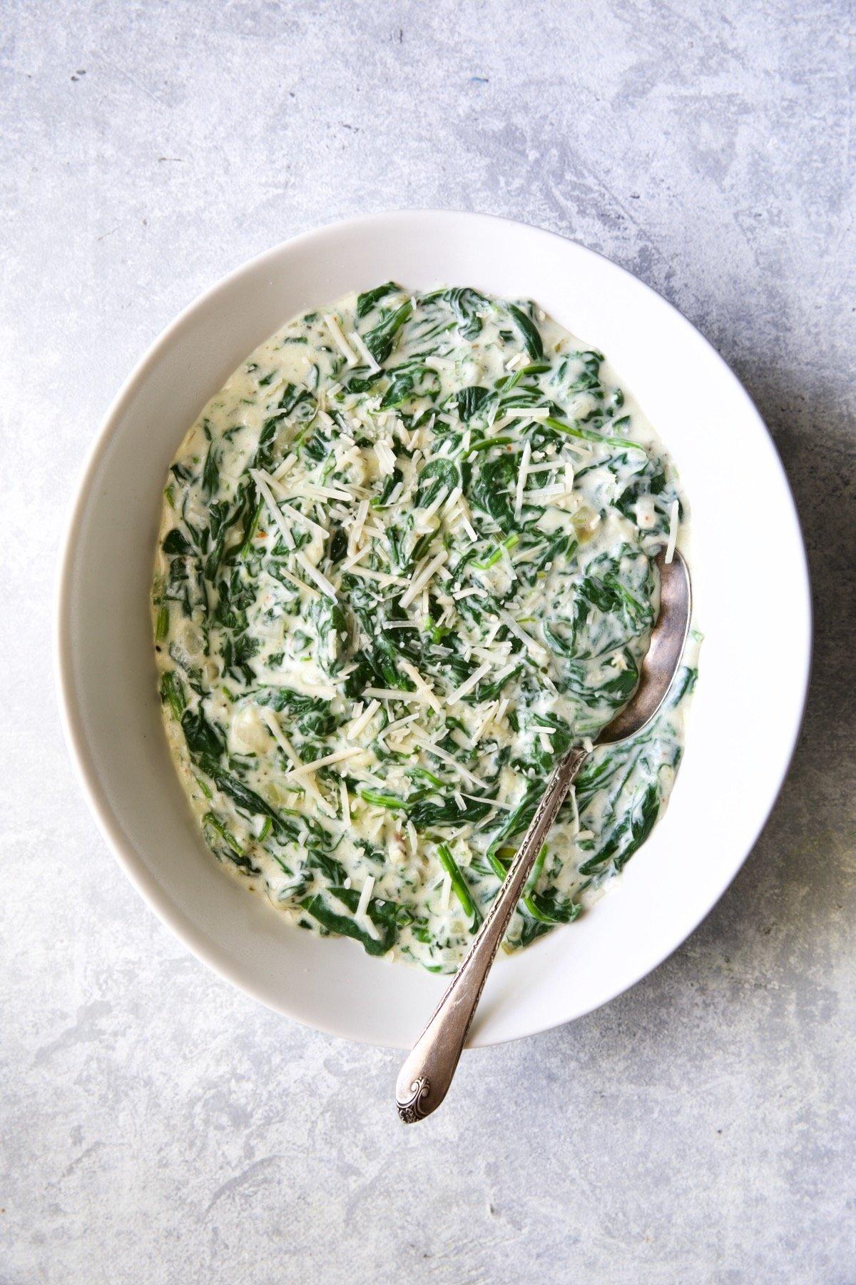 Creamed spinach in a white bowl, garnished with nutmeg, beside a serving spoon, cozy lighting
