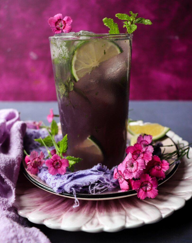 a glass of Violet Drink garnished with fresh berries and mint sprigs, sitting on a sunlit table