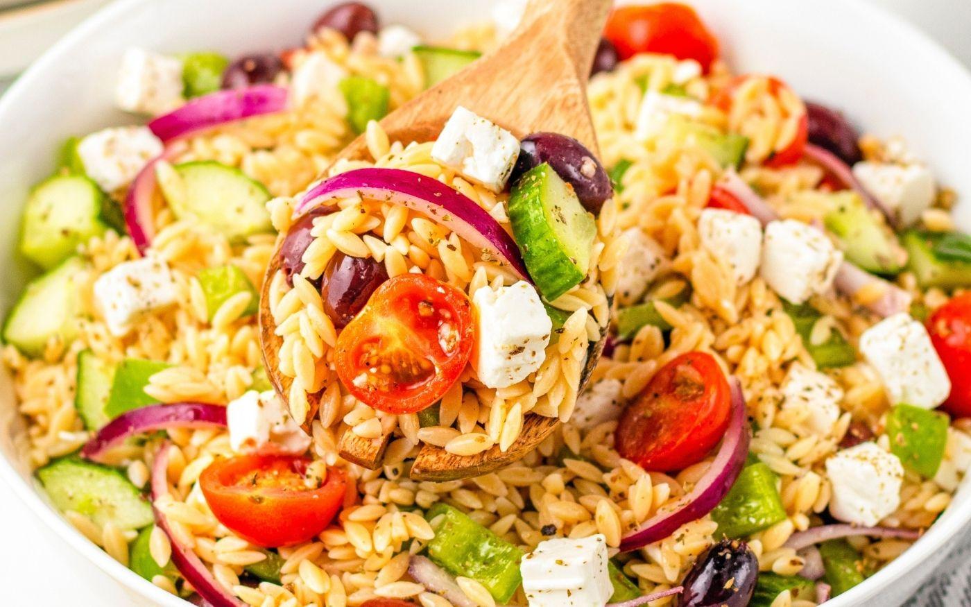 a person tossing Greek orzo pasta salad in a large bowl with a wooden spoon