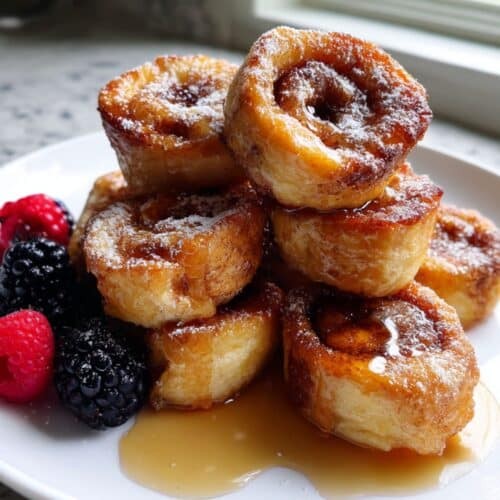 plate of golden brown cinnamon roll french toast bites drizzled with gooey white glaze, with a side of fresh berries and a coffee mug