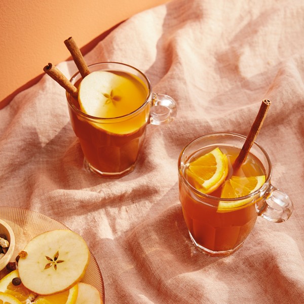 Warm Friendsgiving spiced apple drink in clear mugs with cinnamon sticks and orange slices, cozy autumn setting