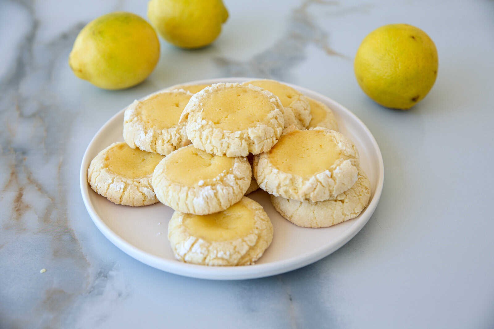 Plate of bright yellow lemon cookies dusted with powdered sugar, fresh lemons, and lemon zest in the background, a cozy kitchen setting.
