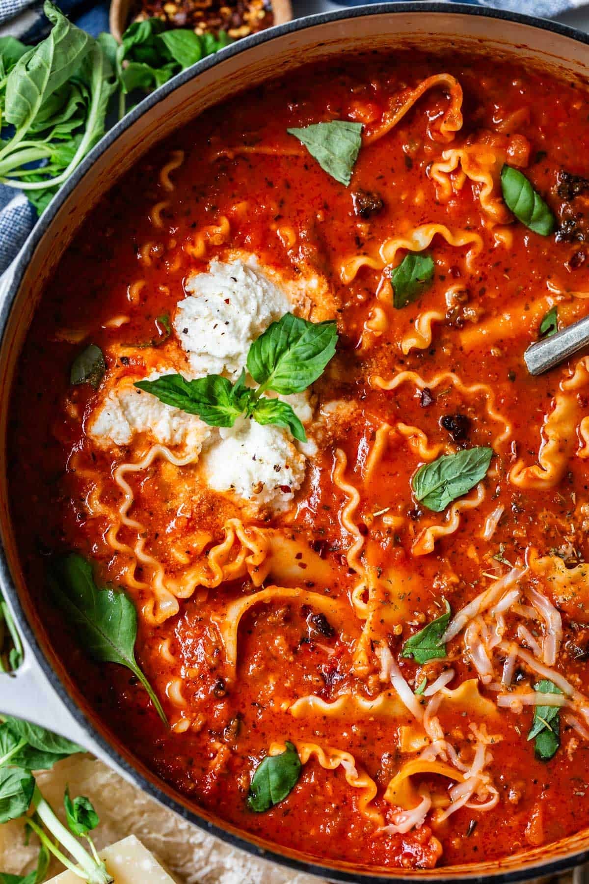 A steaming pot of lasagna soup with a ladle scooping a serving, garnished with fresh basil and ricotta cheese.