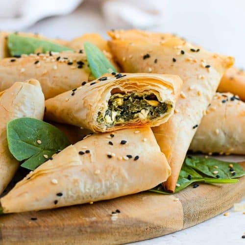 Golden brown spinach feta pastry triangles stacked on a rustic wooden board with fresh herbs
