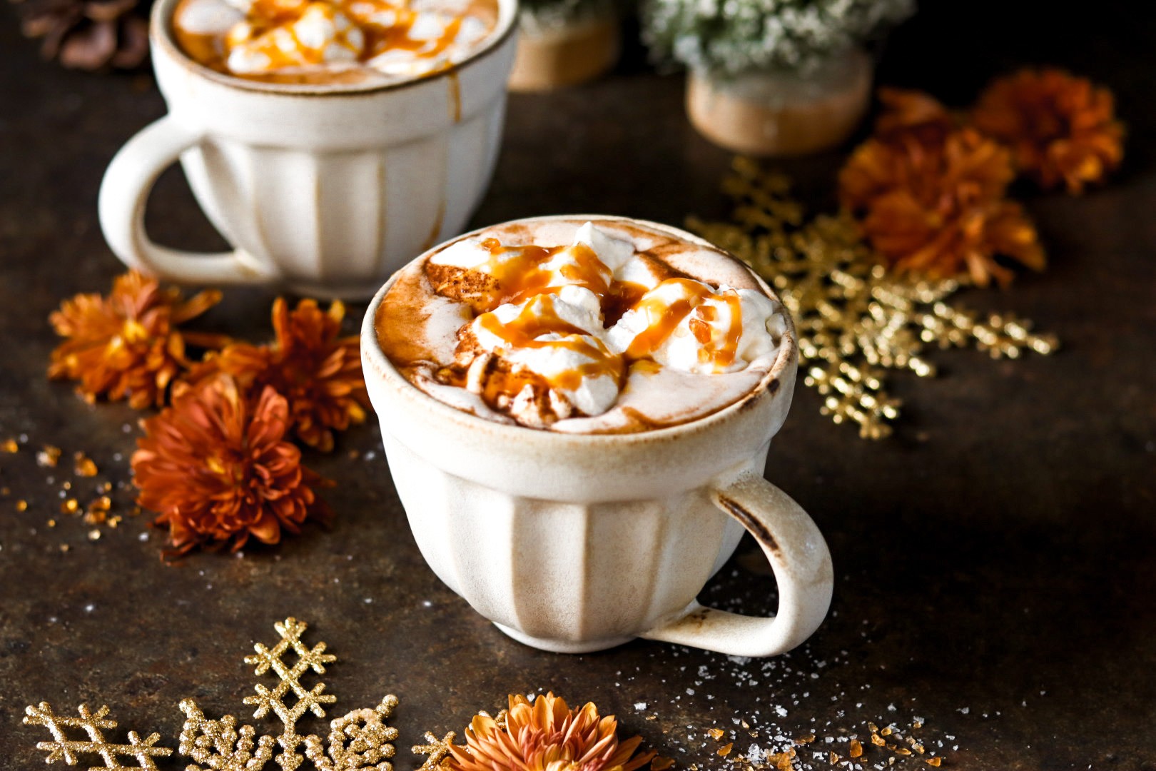 Overhead shot of two mugs of salted caramel hot chocolate with generous whipped cream, caramel drizzle, and a sprinkle of sea salt, on a rustic wooden table with a cozy blanket, a book, and warm lighting