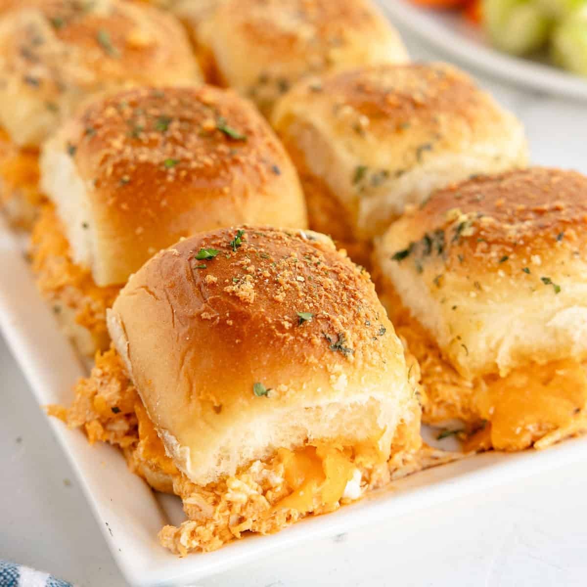 Close-up photo of buffalo chicken sliders on a platter, with a blurred background of a party, celebrating