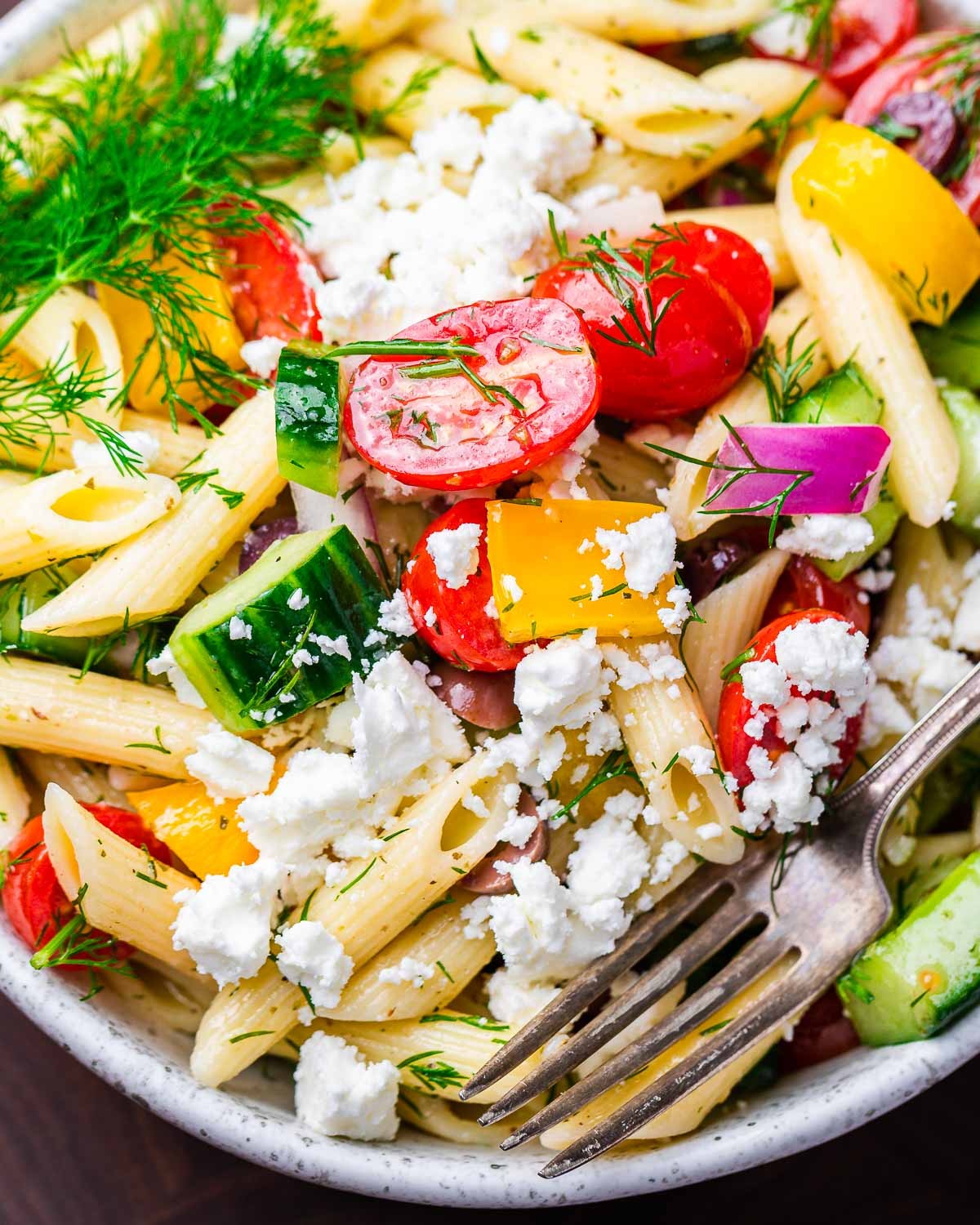 close up shot of vibrant Greek pasta salad with marinated tomatoes in a white bowl, feta cheese, olives, and herbs
