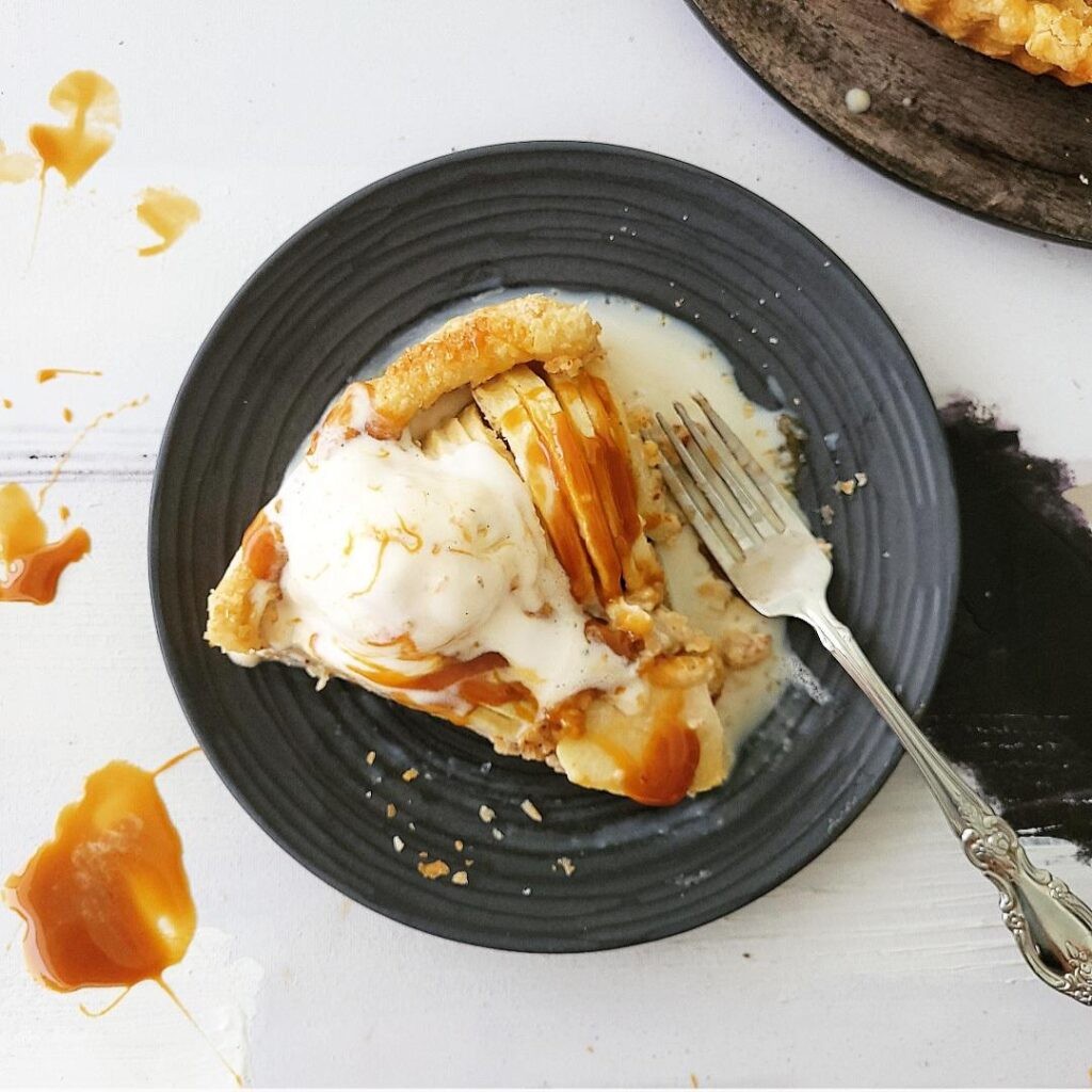 overhead shot of a golden-brown caramel apple tart on a wooden table, a slice removed, with a dollop of vanilla ice cream melting beside it